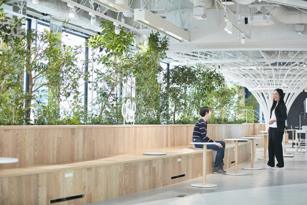 窓際に連なる木製ベンチと樹木の植栽が並ぶオープンスペース(JT本社ワークプレイス「ときの森」)