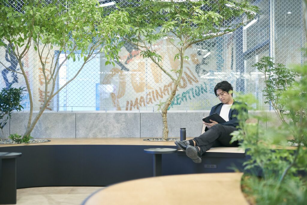 屋内の木陰のベンチに腰かけてタブレットを見る人(TBSホールディングス 本社フリースペース「PARK!」)